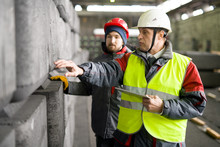Waist Up Portrait Of Mature Worker  Wearing Warm Jacket And Hardhat Using Digital Tablet While Discussing Concrete Blocks Production With Trainee In Workshop