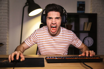 Wall Mural - Portrait of angry irritated gamer guy screaming while playing video games on computer, wearing headphones and using backlit colorful keyboard