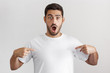 © Damir Khabirov - Indoor photo of handsome Caucasian male isolated on gray background wearing white blank T-shirt and pointing to it with both hands with mouth opened with surprise, amazed by good commercial offer