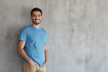 Horizontal portrait of young European male standing isolated against grey textured wall feeling confident and looking optimistic in casual clothes, copyspace for advertising your goods or services