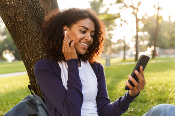 Sticker - Young woman sitting outdoors in park using mobile phone listening music with earphones.