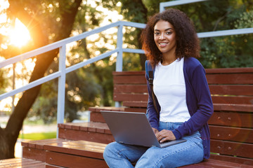 Poster - African young woman sitting outdoors in park on steps using laptop computer.