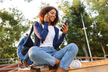 Poster - Woman sitting outdoors in park using mobile phone listening music with earphones.