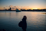 Fototapeta Do akwarium - Traveler man with backpack standing on the sea coast and looking at the bay at sunset. Dusk. Cold winter day in Norway, Oslo