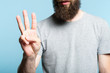 © golubovy - bearded man showing number three with his hand. cropped shot of a male torso on blue background. casual hipster in grey t-shirt counting gesture.