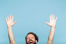Happiness Enjoyment And Laugh. Excited Man With Hands In The Air. Portrait Of A Young Bearded Guy On Blue Background. Emotion Facial Expression. Feelings And People Reaction.