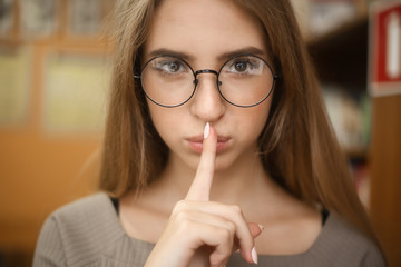 Young student schoolgirl lady with long hair wearing eyeglasses standing in empty classroom looking camera showing silence gesture.