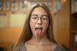 © Sunrise Team - Pretty young student schoolgirl lady with long hair wearing eyeglasses standing in empty classroom looking camera showing tongue and emotions grimace winking.