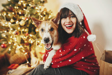Happy Girl In Santa Hat Hugging With Cute Dog On Background Of Golden Beautiful Christmas Tree With Lights In Festive Room. Family Warm Atmospheric Moments. Winter  Holidays