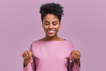 Funny Surprised African American Female Looks With Joyful Eyes Down And Indicates As Shows Something, Sees Comic Things, Isolated Over Lavender Wall. Positive Amazed Young Woman Poses In Studio
