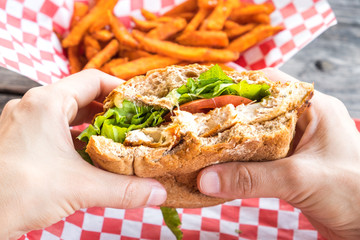 Woman hands hold burger style grilled chicken breast sandwich with lettuce and tomatoes and a side of sweet potato fries