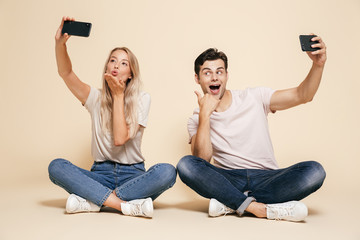 Poster - Young loving couple sitting isolated over beige wall background make selfie by mobile phones.