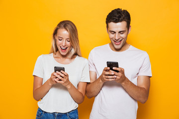 Wall Mural - Happy excited young loving couple standing isolated over yellow wall background using mobile phones.