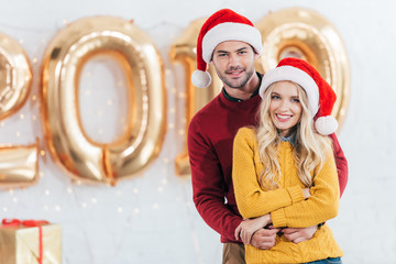 Wall Mural - beautiful smiling couple in santa hats hugging at home with 2019 new year golden balloons