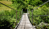 Fototapeta Mosty linowy / wiszący - Wooden hanging bridge leading forward over green mountain river.