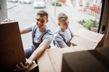 Two young handsome smiling movers wearing uniforms are unloading