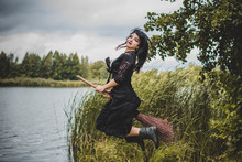 Young Beautiful And Mysterious Witch Woman Flying On The Broom In Woods. Levitation, A Witch In A Hat Flies On A Broom, Free Space. Halloween Holiday, Magic