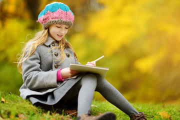 Cute little girl sketching outside on beautiful autumn day. Happy child playing in autumn park. Kid drawing with colourful pencils.