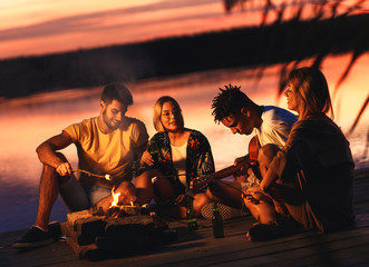 Group of young friends enjoying at the lake at night. They sitting around the fire singing and having fun.