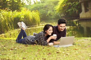 Wall Mural - Attractive asian couple watching movies on laptop while lying on the green grass