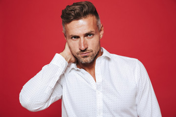 Portrait of masculine serious man 30s with stubble in white shirt looking on camera, isolated over red background