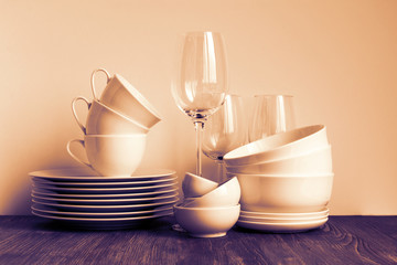 Wall Mural - White dishware stacked on a wooden table against white background with transparent wineglasses. Toned.