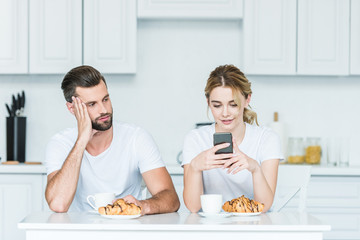 Wall Mural - bored man looking at smiling girlfriend using smartphone during breakfast