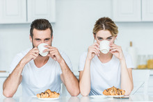 Young Couple Drinking Coffee And Looking At Camera While Having Breakfast With Coffee And Croissants