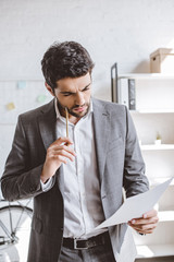 Wall Mural - pensive businessman reading documents and touching cheek with pencil in office