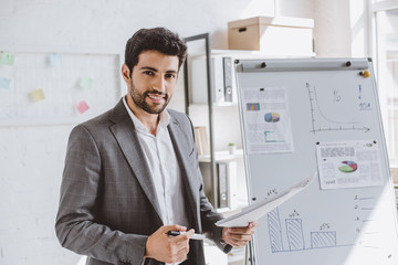 Wall Mural - smiling handsome businessman presenting project on flipchart and looking at camera in office