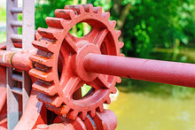 Close-up Of A Red Metal Gear On The Floodgate. Mechanism To Open The Dam. Old Toothed Wheel On The River. Transfer To The Opening Of The Dam. Old Cogwheel