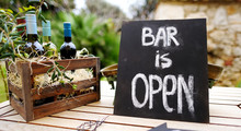 "Bar Is Open" Sign And Vintage Wooden Crate Full Of Wine Bottles Decorated With Olive Branches On A Table.