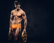© Fxquadro - Handsome naked ectomorph bodybuilder with stylish hair in underpants posing with a barbell disk on a dark background.