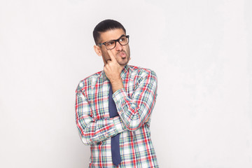 Wall Mural - Portrait of confused handsome bearded businessman in colorful checkered shirt, blue tie and eyeglasses standing touching face and looking away. indoor studio shot, isolated on light grey background.