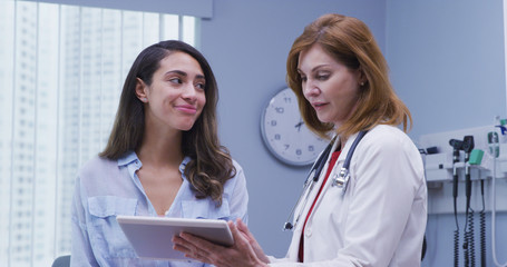 Wall Mural - Portrait of beautiful young latina patient consulting with doctor about test results. Close up of pretty hispanic woman looking at tablet device with doctor indoors medical clinic