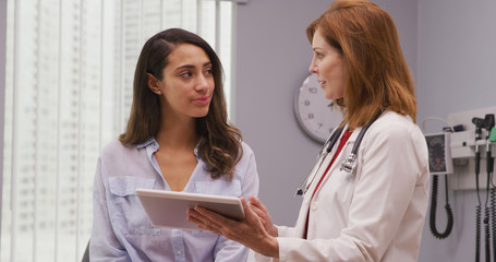 Wall Mural - Portrait of lovely young hispanic woman meeting with senior doctor about pregnancy results. Close up of mid aged caucasian doctor using portable tablet to review health history with latina patient