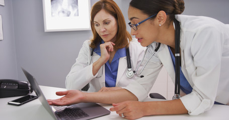 Wall Mural - Intelligent young latina doctor looking at laptop computer and talking with elder physician. Two medical doctors using notebook computer to discuss patients healh condition