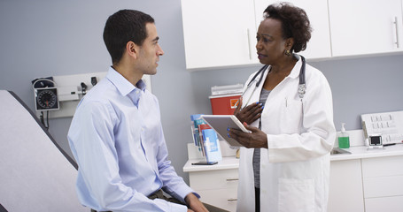 Wall Mural - African senior medical doctor using high-tech elctronic notebook to take notes of patient. Portrait of young hispanic patient consulting with middle aged black doctor indoors medical clinic