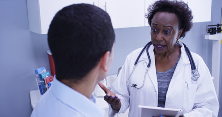 Close view of black medical doctor discussing health issues with young male patient. Senior African doctor using portable tablet to take notes of patients health