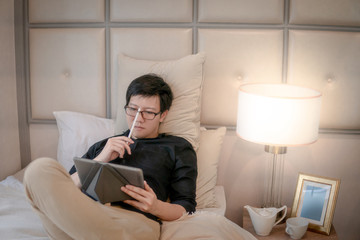 Young Asian business man using digital tablet and pen while lying on bed in cozy bedroom. Home living lifestyle with modern electronic gadget concept