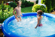 Two Cheerful Cute Little Sisters Playing And Having Fun, Splashing And Jumping In Inflatable Pool At Backyard