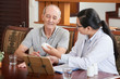 © DragonImages - Woman at table with tablet and papers and showing pills in jar to senior patient sitting near
