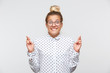 Portrait of happy excited young woman with bun wears polka dot shirt and spectacles keeps fingers crossed, biting lip and making a wish isolated over white background