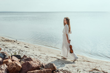 beautiful elegant woman in white dress holding violin and walking on beach near sea