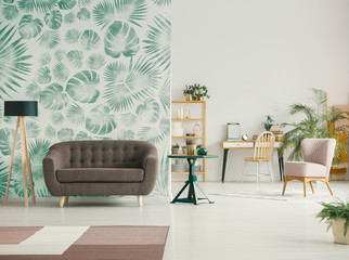 Brown sofa next to lamp in apartment interior with leaves wallpaper and pink chair. Real photo