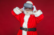 © satyrenko - Christmas. Santa Claus in black virtual reality glasses makes gestures with his hands. Surprise, emotion. New technology. Isolated on red background.