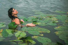 Beautiful Dark Haired Girl In White Dress Posing In River With Water Lilies. Fairytale Story About Modern Ophelia  