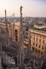 Vertical: Rooftop view of cathedral spires & sculptures, Galleria Vittorio Emanuele II, and distant modern buildings at sunset from Duomo di Milano.