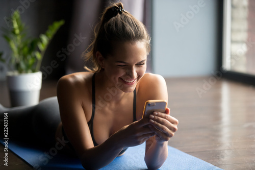 Young sporty woman after practicing yoga, break in doing exercise, relaxing on yoga mat, texting on the cell phone, holding smartphone, friendly smiling reading message, indoor close up, yoga studio