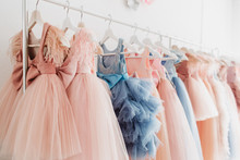 Beautiful Dressy Lush Pink And Blue Dresses For Girls On Hangers At The Background Of White Wall. Kids Dresses With Feathers For Prom And Holiday.